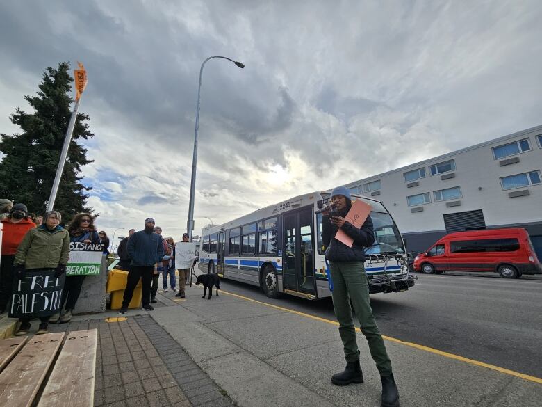 Person speaking into a microphone, bus in the background, people gathered around.