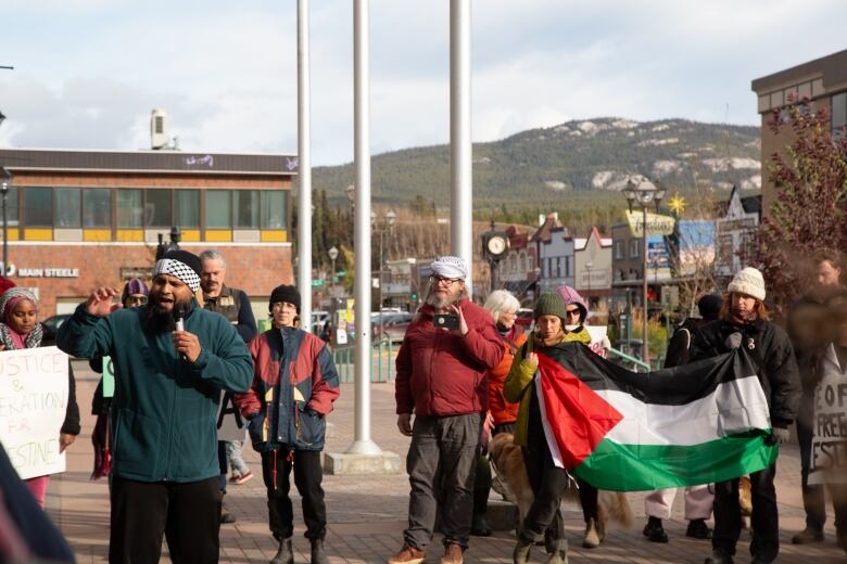 Person speaking to a crowd, Palestinian flag in the background. 