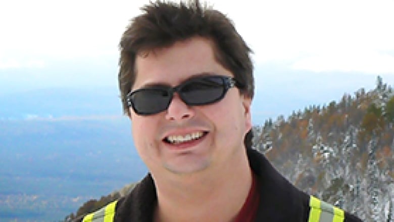 A man in a yellow vest smiling in front of mountains. 