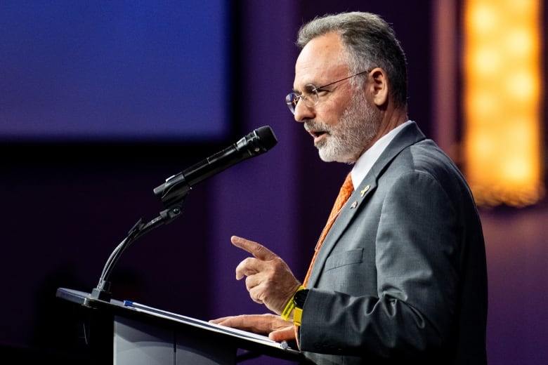 A man in a gray suit points as he speaks into a microphone.