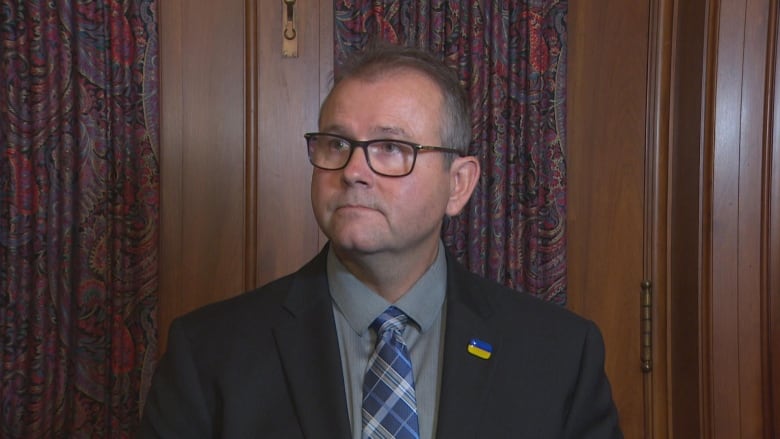 A man in a black suit, grey shirt and striped blue tie looks on.