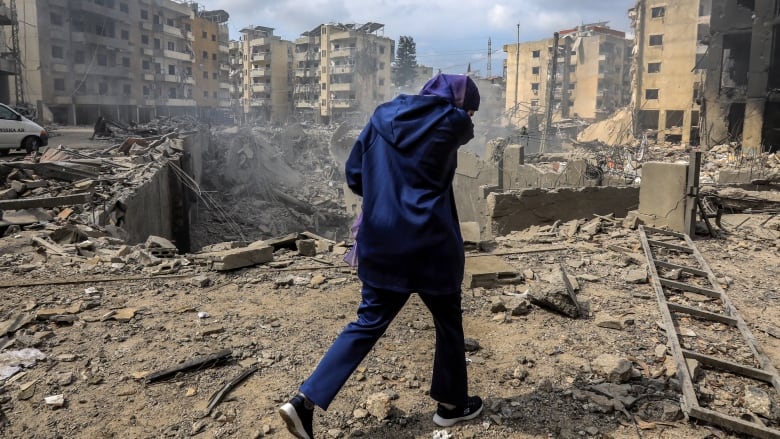 A woman in a jacket and sneakers with her back to the camera walks on dirt ground strewn with debris, as a large crater and heavily damaged multistorey buildings are shown.