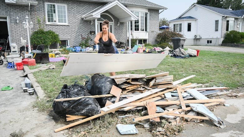 Woman holds a piece of drywall above a pile of wood, garbage bags and other rubble in front of a house.