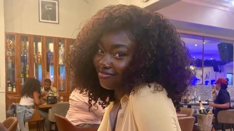 A young Black woman wearing a tan long sleeve button-up sitting down at a restaurant table. She has her hair out and is smiling at the camera. 