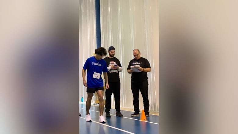 A young man in a blue t-shirt on a track with two men nearby holding notepads.