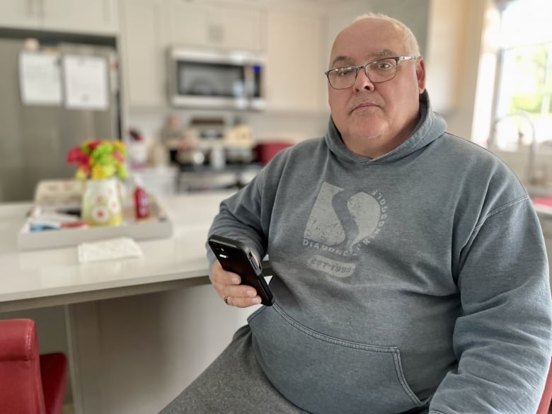 Garth Jay sits in his kitchen, holds a phone, and stairs at the camera. 