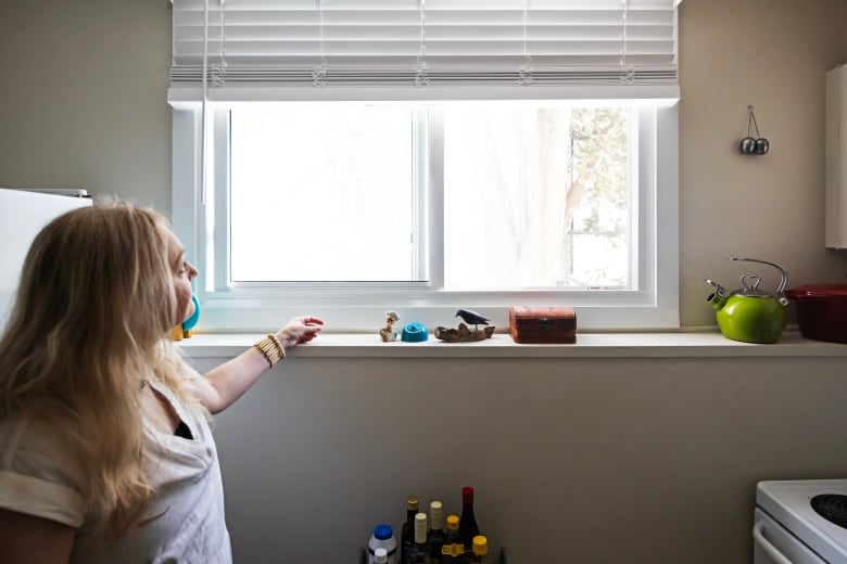 A woman stands near a kitchen window that has a white frame, her arm resting on the edge of the window. 