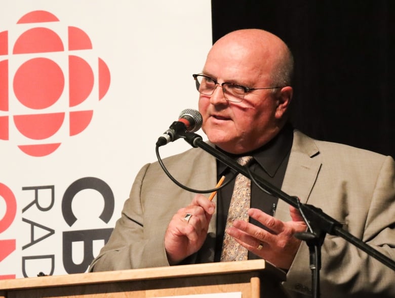 A man in a tan suit with a black shirt and light patterned tie wearing glasses uses a pencil to count on his hand.