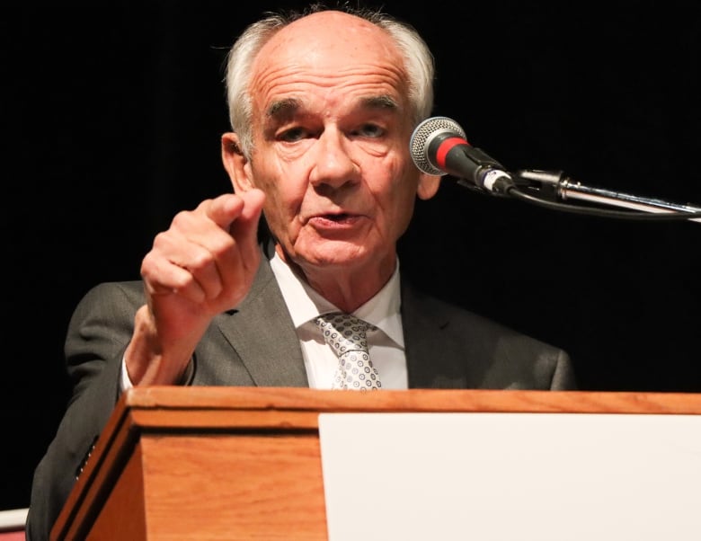 A man in a grey suit with white shirt and silver patterned tie points his finger and speaks into a microphone.