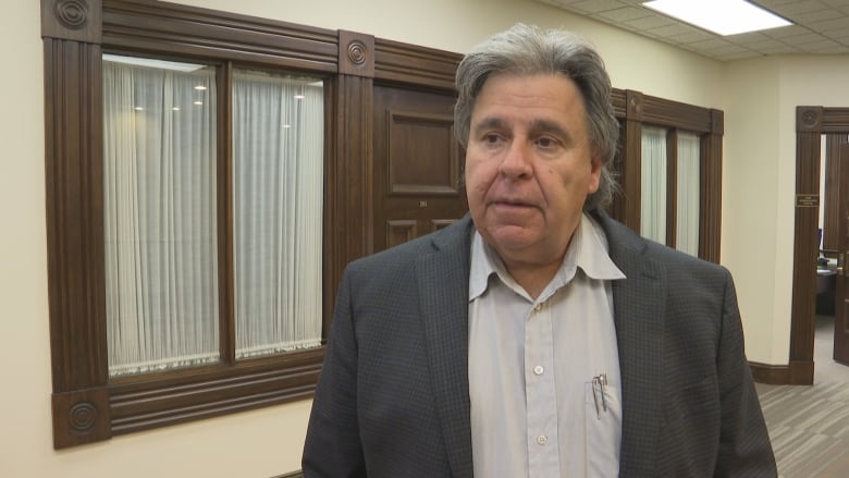 A man in a grey suit is doing an interview. He is framed from the chest up. His face is serious. There in one CBC microphone in front of him 