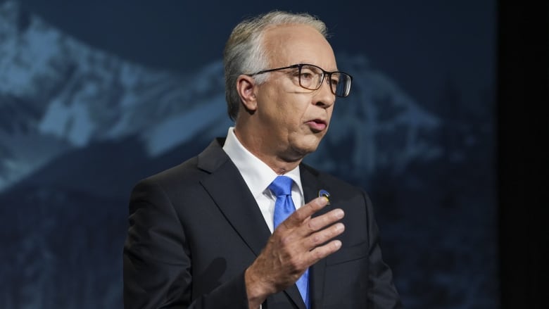 A man looks to his left as he speaks in front of a mountain backdrop.