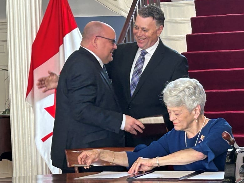 Dennis King pats Gilles Arsenault on the back while Lieutentant-Governor Antoinette Perry, seated, smiles and sorts through papers.