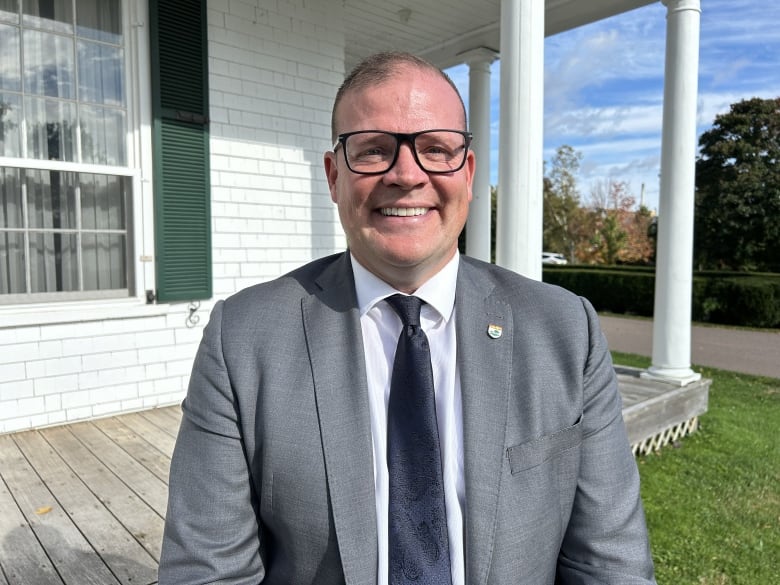 A smiling man with glasses in a grey suit and dark blue tie stands in front of a white-painted colonial-style verandah.