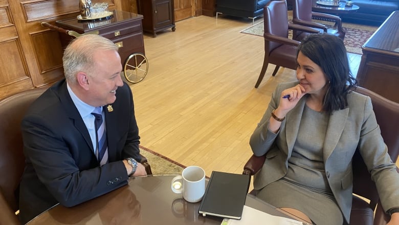 Premier Danielle Smith, on the right, sits leaning back from an office table. On her left, a man in a suit looks at her and smiles. 