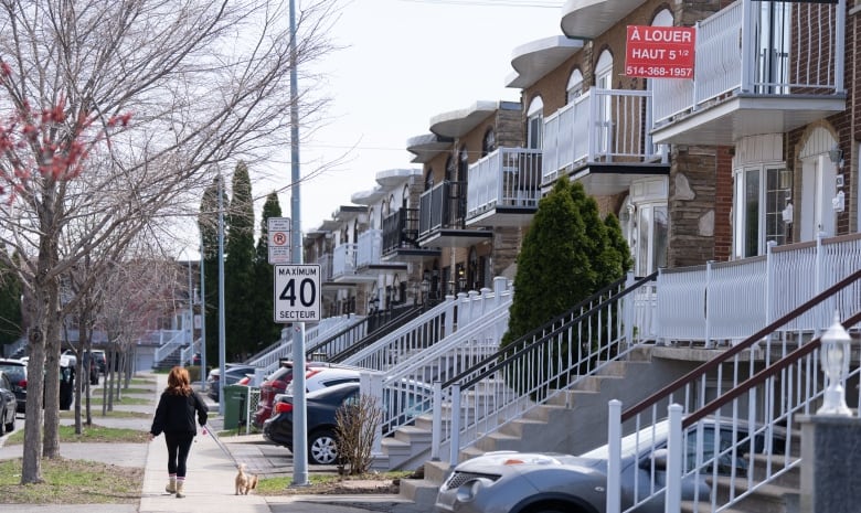 A residential street is shown, with a person walking down the sidewalk. Trees are bare of leaves. A red sign hangs outside of the second floor of a house, attached to the balconey. 