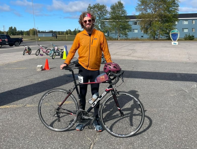 A smiling man wearing an orange jacket stands with a bicycle.