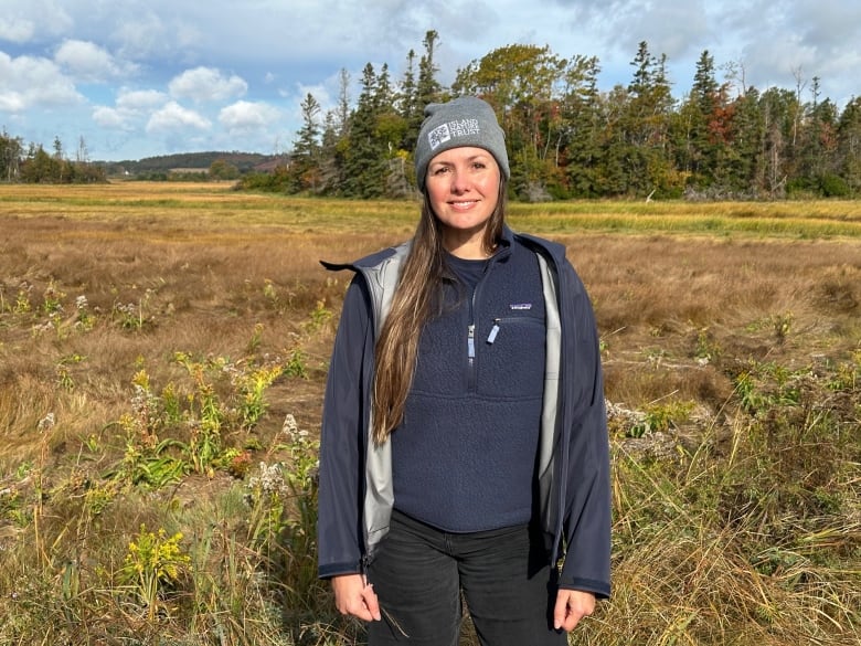 A woman wearing a blue jacket and grey toque stands near a marsh 