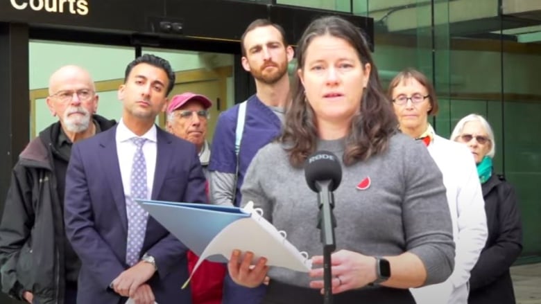 A woman speaks at microphone outside of a courthouse, with a small group of people standing behind her. 