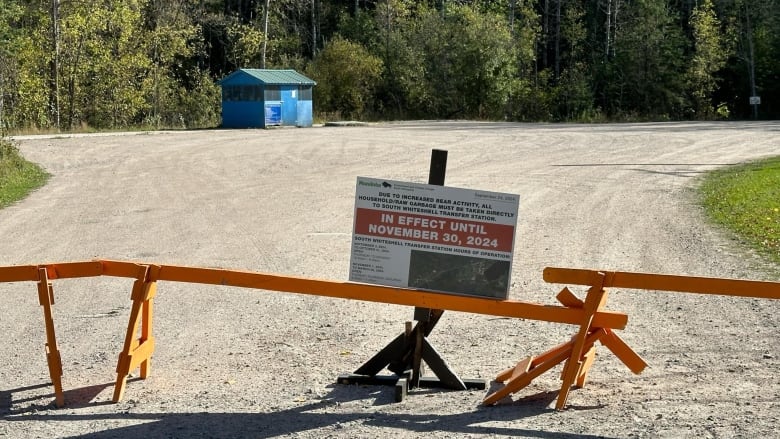 A sign is perched in a gravel parking lot with orange barricades preventing people from going through. 