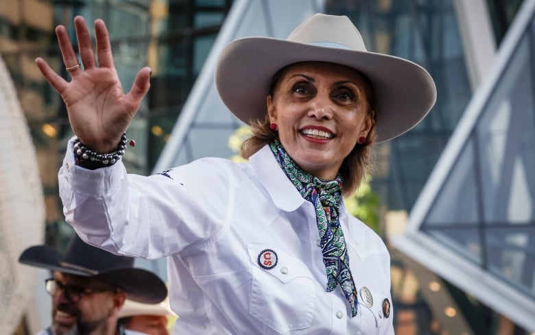 A woman wearing a cowboy hat waves to the camera.