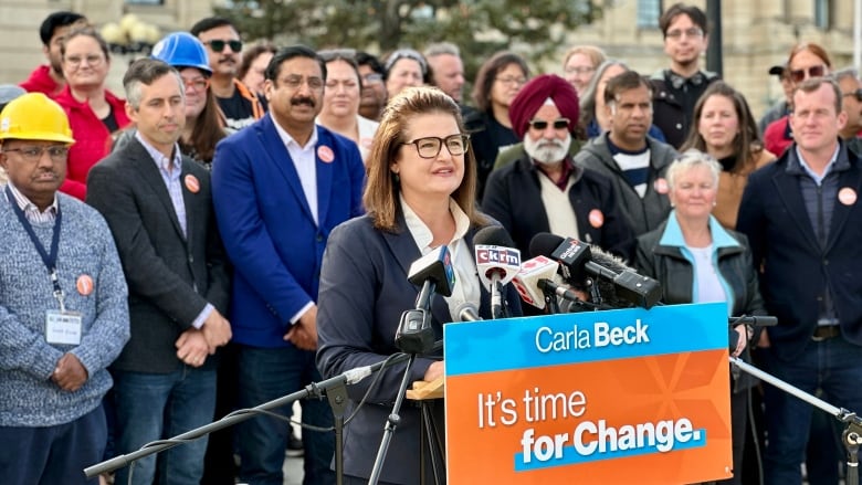 A woman stands behind a podium. The woman is wearing a white button up shirt and a blue jacket. A group of people stand behind her. 