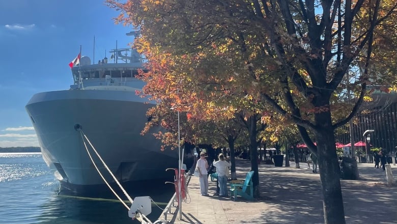 The stern of a large navy patrol ship is visible, docked and tied to the shore in TOronto Harbour. Two people stand beside it on a sunny fall day