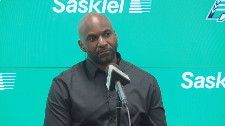 A Black man wearing a black button up shirt and sitting in front a microphone in front of a green background with the Saskatchewan Roughrider's logo. 