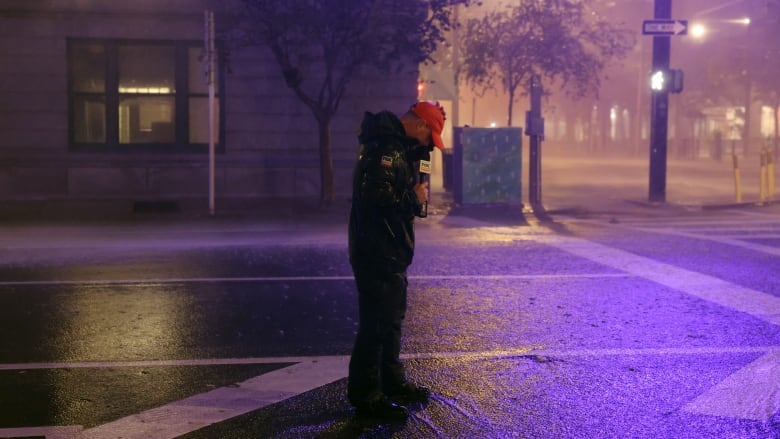 A man in a rain jacket and red ball cap holds a microphone and stands in the middle of the street in the rain.