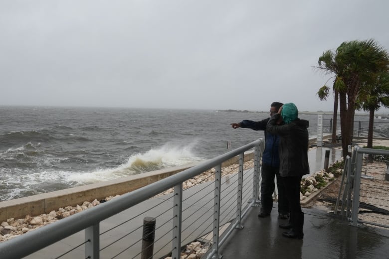 Two people in rain gear stand on a pier as winds whip the water into waves. 