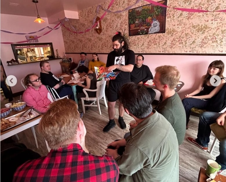 A room with a group of people sitting on chairs. One person is standing and holding a book.