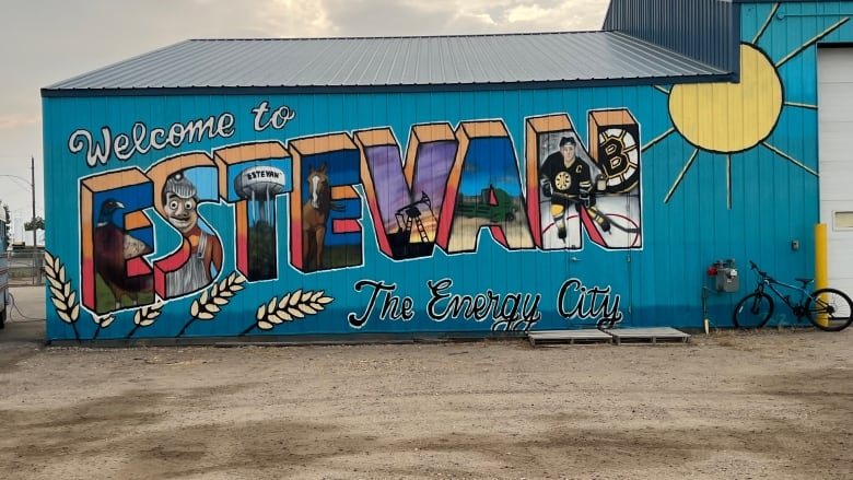 A painted sign on a building reads 'Welcome to Estevan, The Energy City,' with the letters featuring a painting of a duck, a person, a water tower, a horse, a pump jack, agricultural equipment and a hockey player.