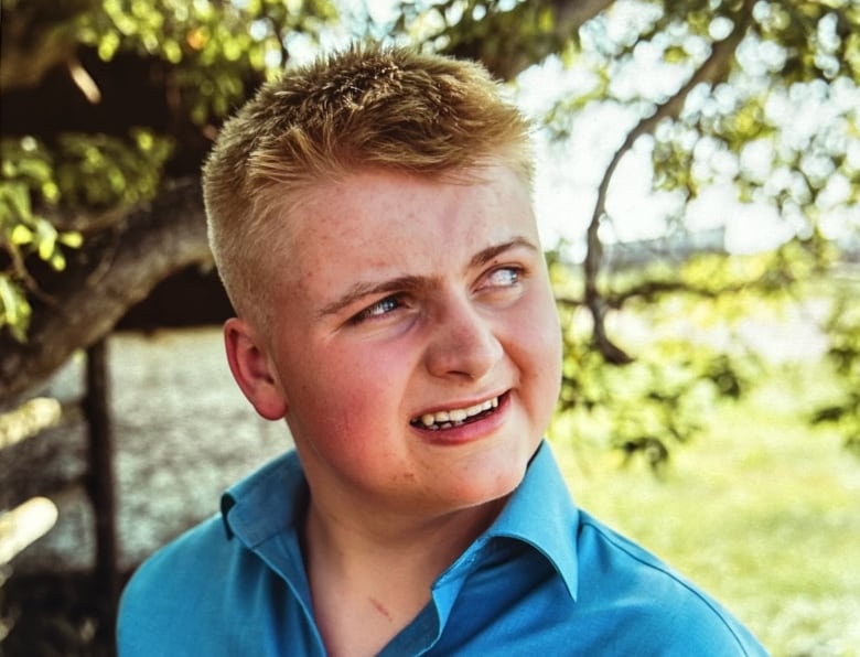 A young blonde man in a blue shirt looks to the side, with trees and greenery seen behind him.