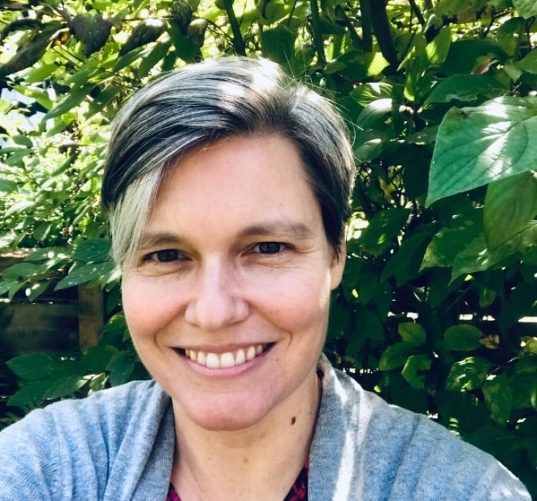 A woman with short hair poses for a selfie with leafy shrubbery in the background.