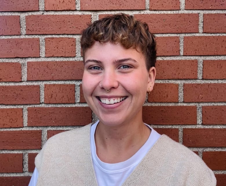 A smiling person, wearing a beige vest, in front of a brick wall.