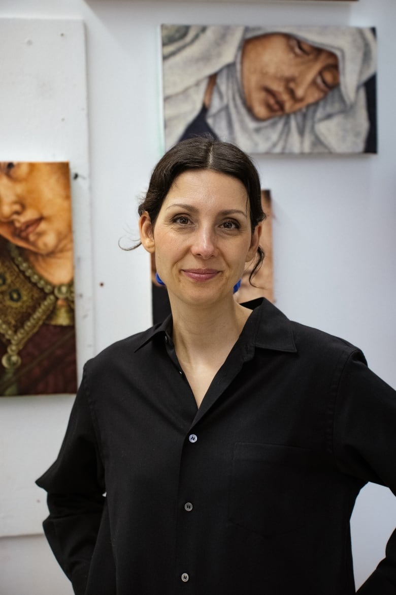 A woman with dark hair and a dark shirt stands in front of a wall of paintings. 