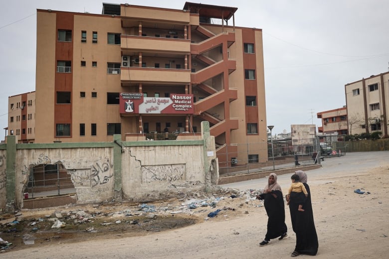 Two women, one holding a child in her arms, walk past a damaged wall in front of a large building with a sign reading Nasser Medical Complex in English and Arabic. 