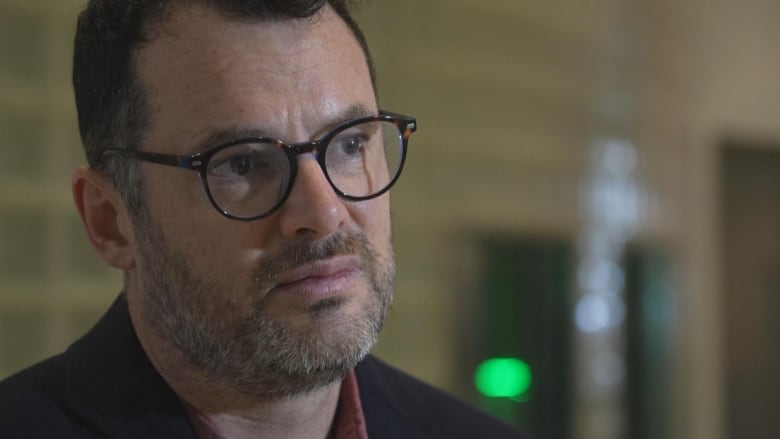 A close up shot of a man with a beard dark hair and glasses siting in an office.   