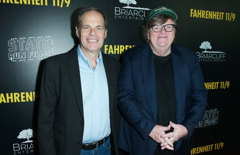 Two men wearing blazers, one with a baseball hat, pose for a photo. 