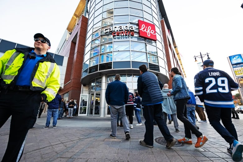 A person in uniform in front of the Canada Life Centre. People, some wearing hockey jerseys, are walking toward the building's entrance.