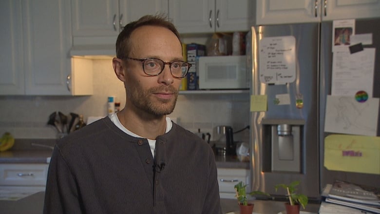 A man in classes stands in a kitchen. There are small plants on the island and children's drawings on the fridge behind him.