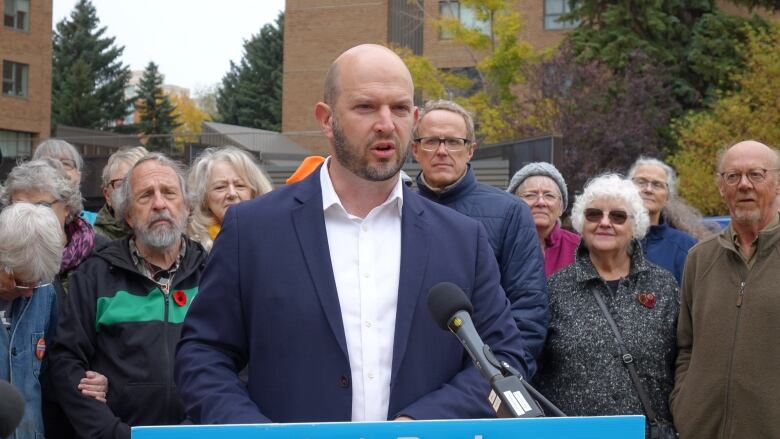 A man in suit speaks at a podium outside with several others standing behind him.