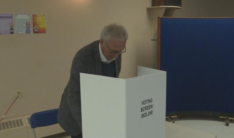 A man with thinning white hair and wearing a grey suit jacket leans over a table where a white cardboard is set up that says Voting Screen.