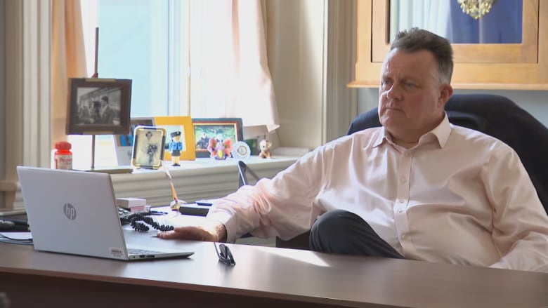 A man sits inside an office. He wears a salmon coloured oxford shirt. A desk is in front of him. A laptop and sunglasses can be seen on the desk