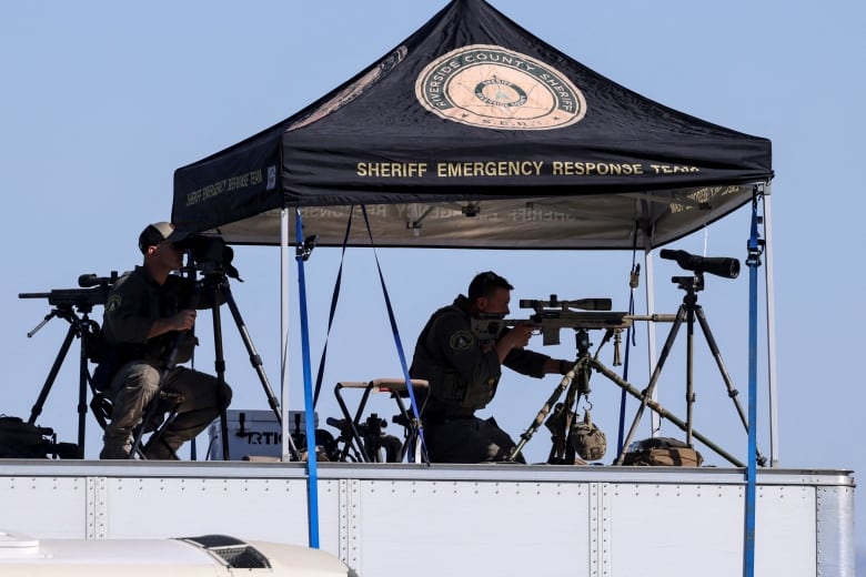 Law enforcement personnel look through binoculars and scoped rifles while situated atop a truck and under a tent.