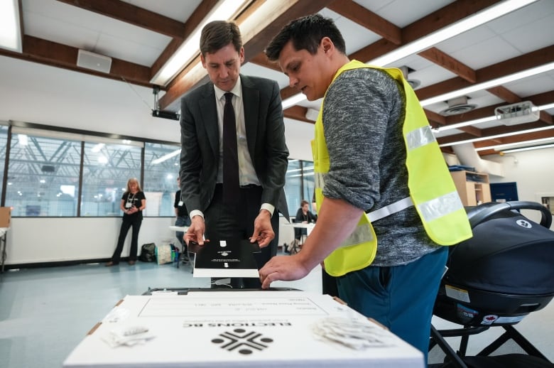 A tall man in a grey suit places an election ballot on a table while a man in a high visibility safety vest assists him.