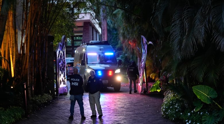 A dark lane in seen at night or evening, lit by the flashing lights of what appears to be a police van. Two people stand in front of the vehicle. Shadowy foliage surrounds the lane on both sides. 