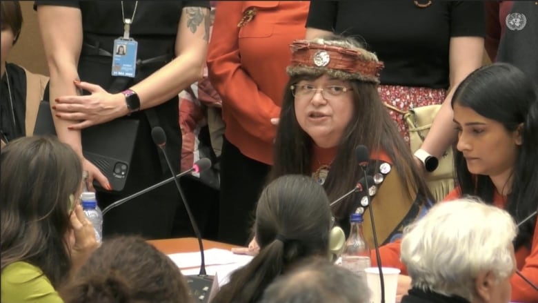 An Indigenous woman wearing traditional regalia speaks in a packed room.