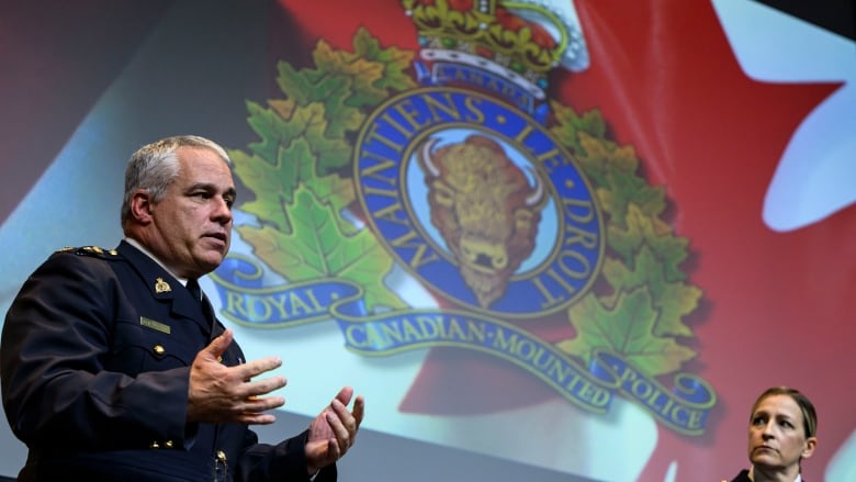 A police officer is pictured with the RCMP logo in the background on a screen as another police officer looks on.