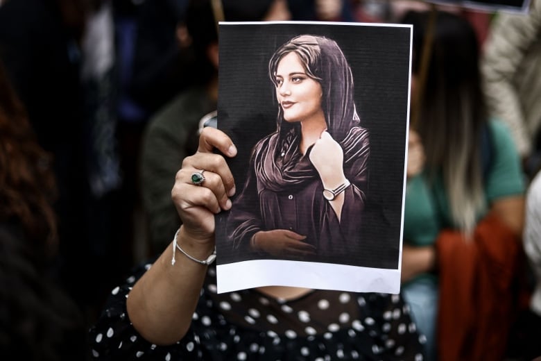 A woman holds a printed photo directly in front of her face showing a portrait of a smiling young woman in a headscarf, pulled slightly back to reveal her long, dark hair.