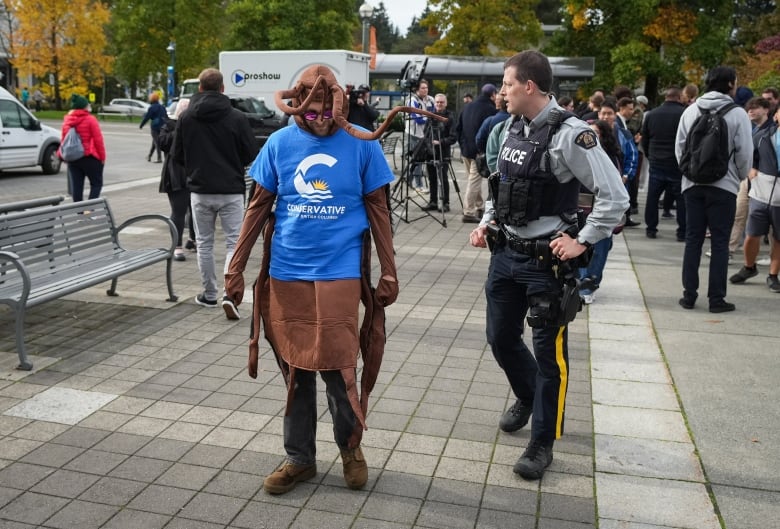 A man stands dressed as an insect wearing a BC Conservatives T-shirt while a police officer speaks to him. 